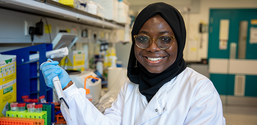 Aisha Yusuf, a member of the Fitzgerald Lab in a white coat, holding an electronic pipette